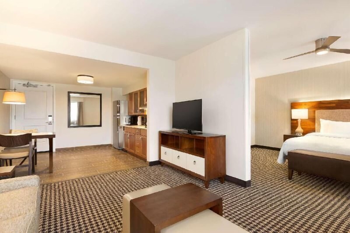 Una habitación de hotel con una cama, un televisor sobre un mueble de madera, un escritorio con una silla, una mesita y una alfombra estampada.