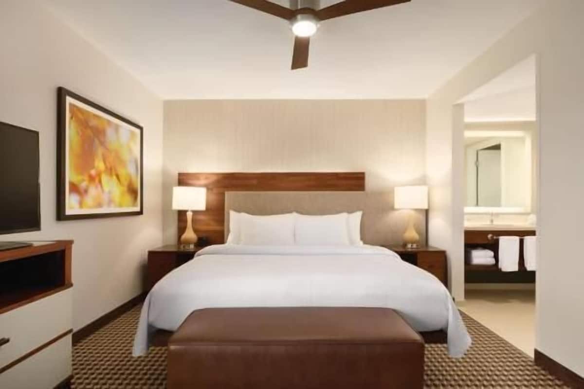 A hotel room with a bed, two bedside lamps, a TV, a painting, a wooden headboard, a bench, and a bathroom with a mirror and towel rack.