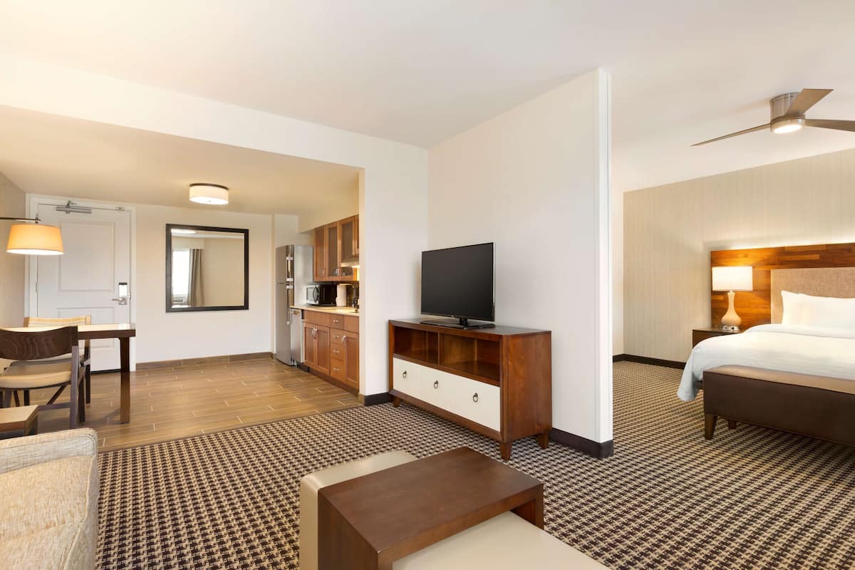 A hotel room with a bed, a TV, a desk, a chair, a sofa, a patterned rug, and a kitchen area in the background.