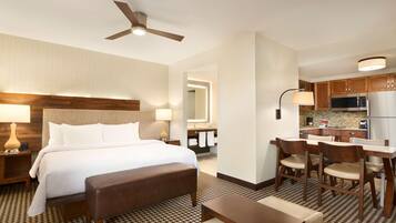 A hotel room with a bed, a desk, chairs, a kitchenette, and a ceiling fan.