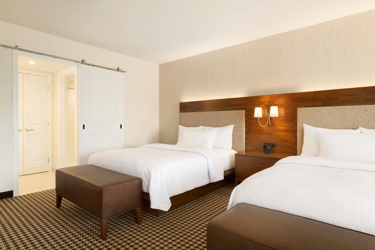 A hotel room with two beds, a bench, a wooden headboard, and a wall-mounted light fixture.