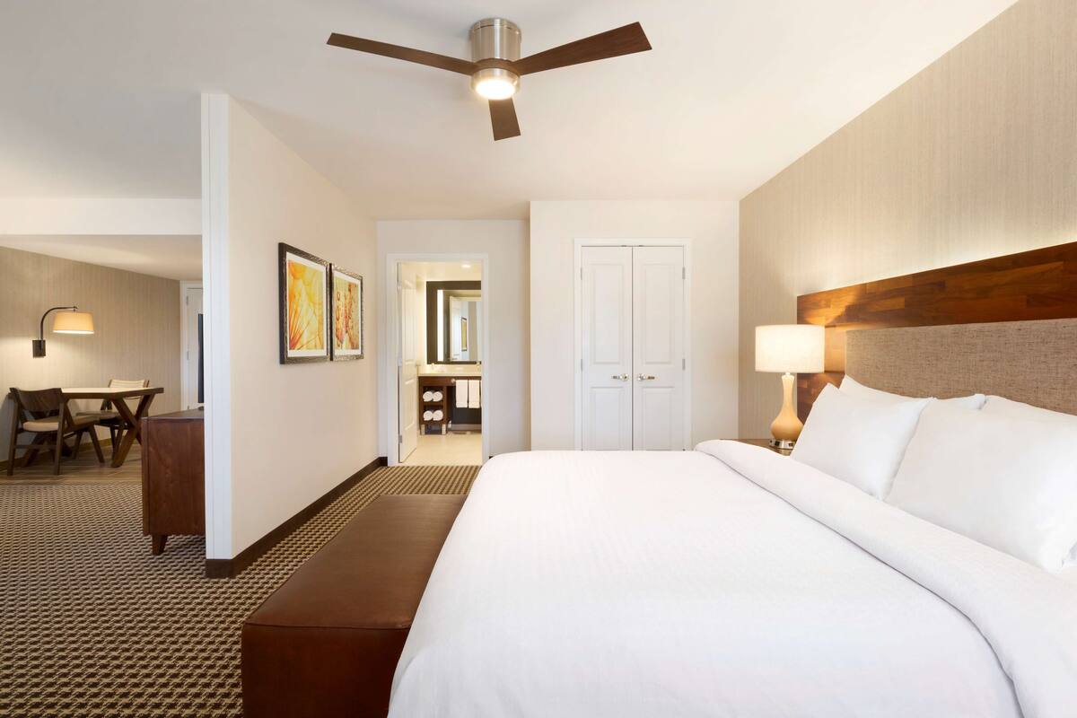 A hotel room with a large bed, a desk, a chair, a ceiling fan, and a bathroom visible through an open door.