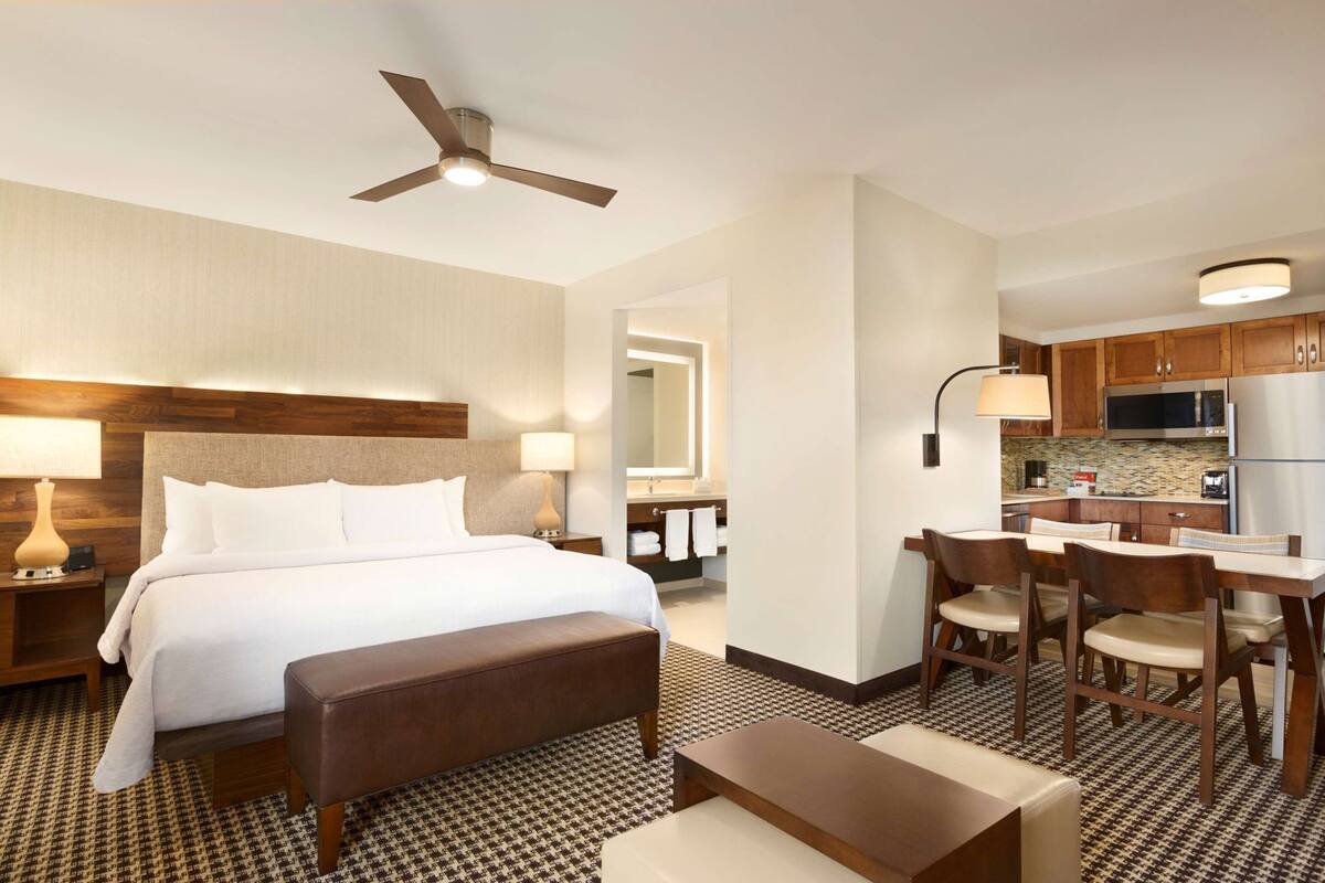A hotel room with a bed, bedside tables, a desk with chairs, a kitchenette, and a ceiling fan.