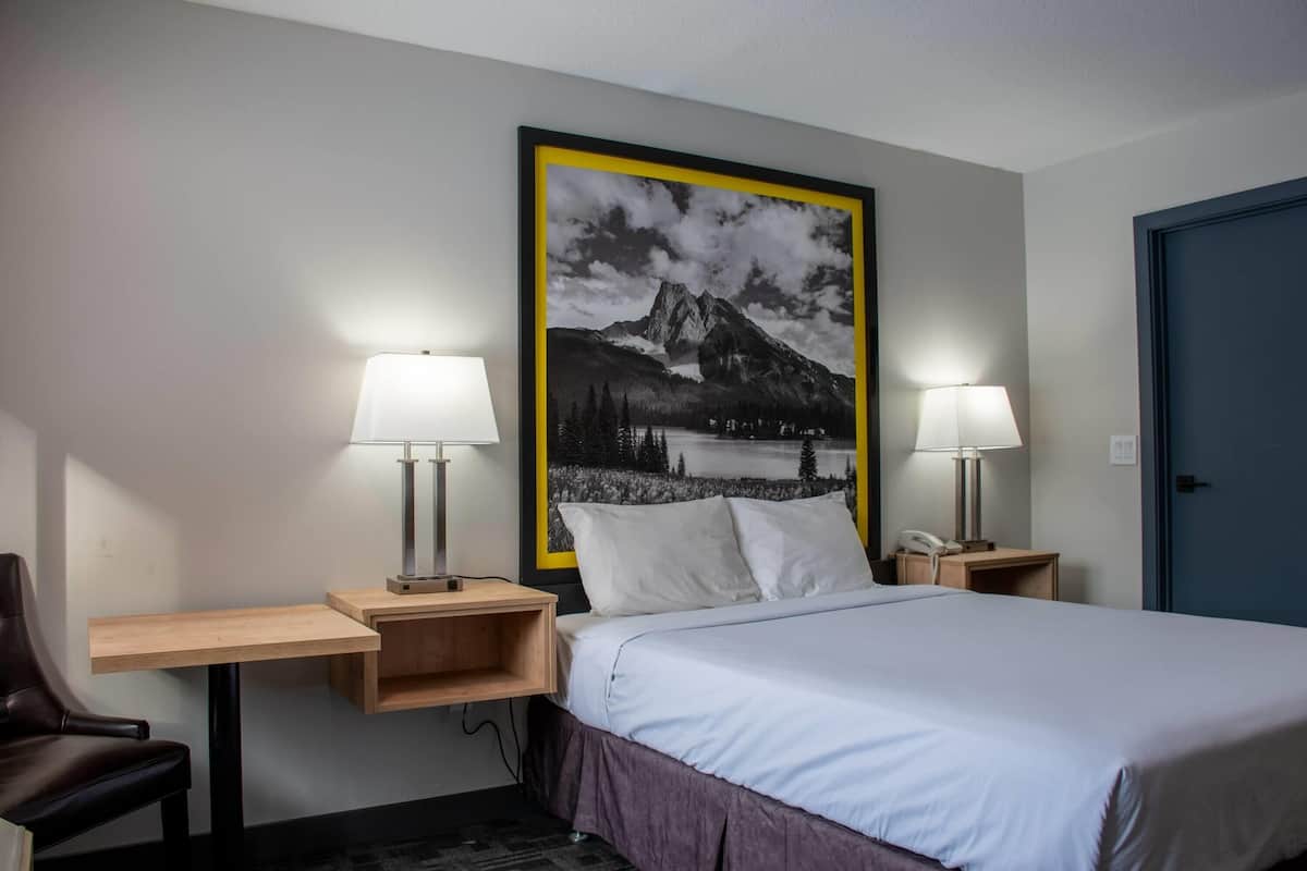 A hotel room with a bed, two bedside lamps, a desk with a chair, a telephone, and a large framed picture on the wall.