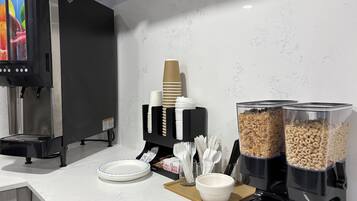 A self-service breakfast station with a coffee machine, paper cups, bowls, and containers of cereal and granola.