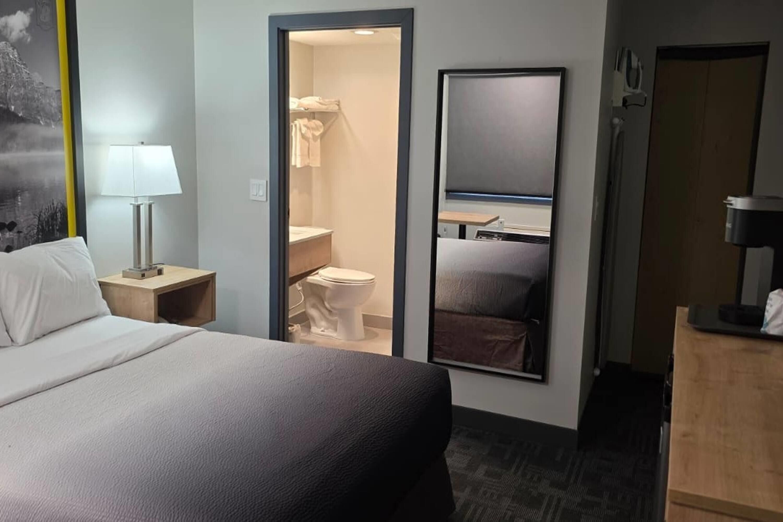 A hotel room with a bed, a nightstand, a mirror, a bathroom with a toilet and sink, and a wooden dresser.