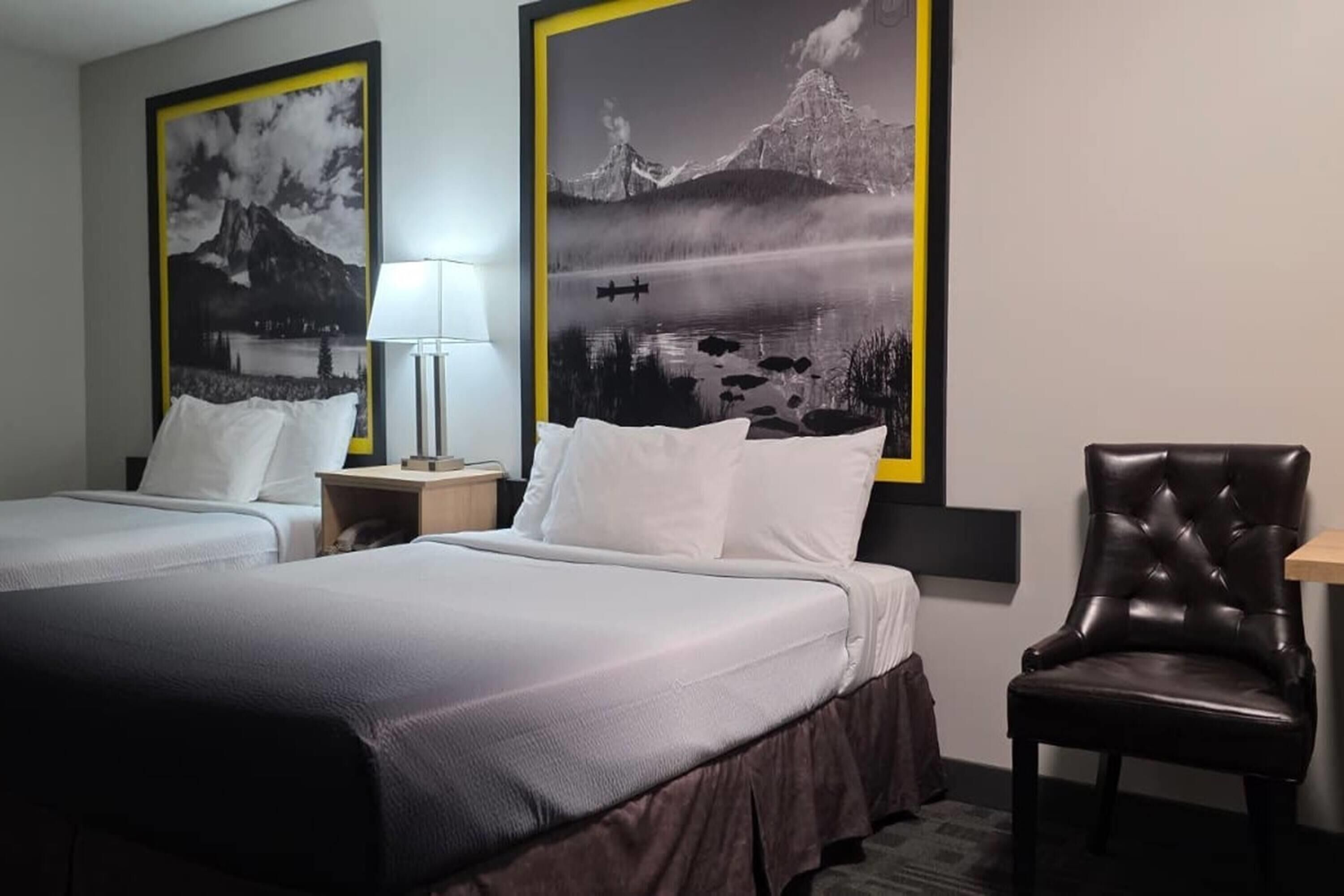 A hotel room with two beds, a chair, a nightstand with a lamp, and two framed pictures on the wall.