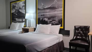 A hotel room with two beds, a chair, a nightstand with a lamp, and two framed pictures on the wall.