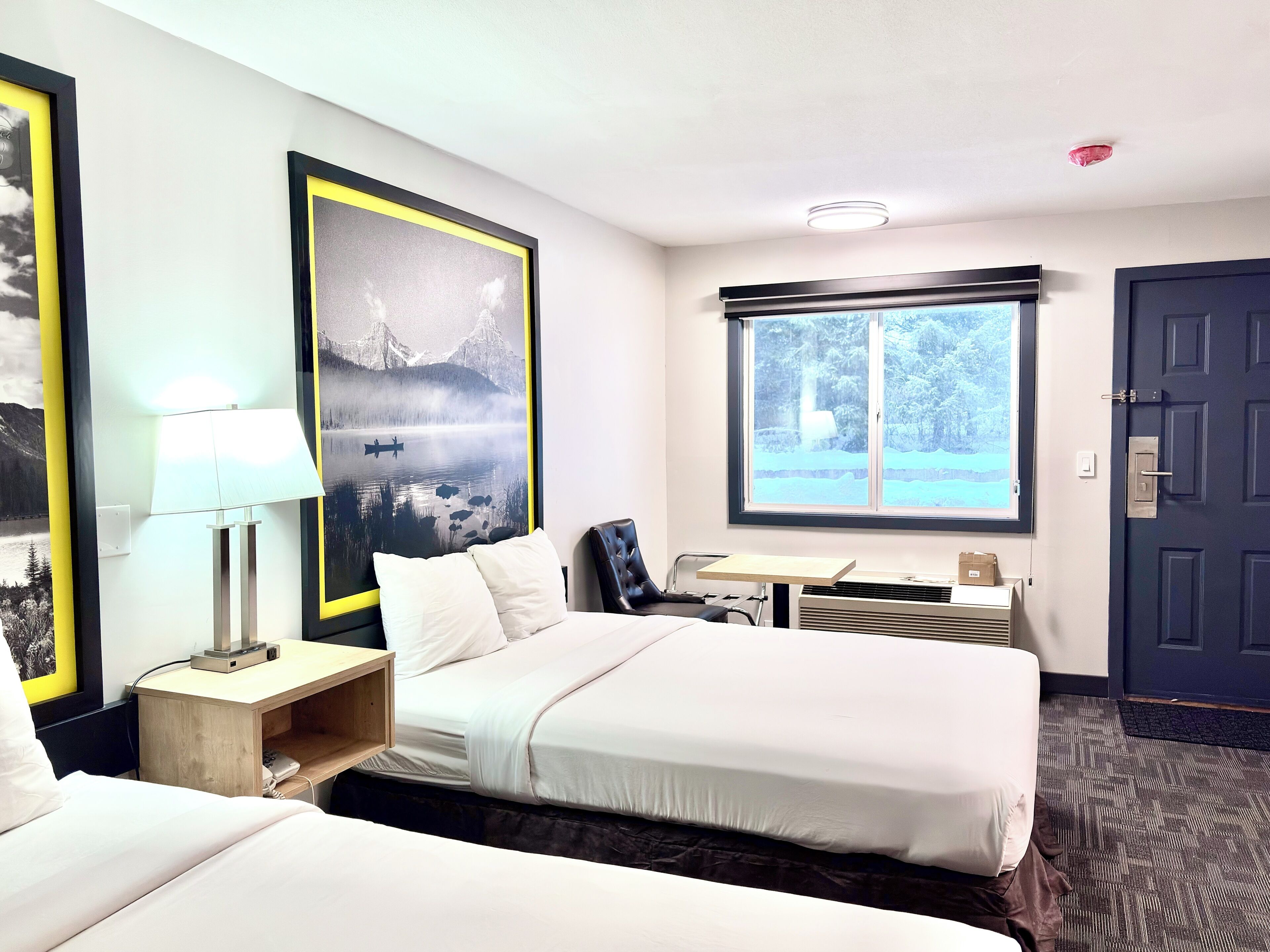 A hotel room with two beds, a desk, a chair, a lamp, a window with a view, and a door.