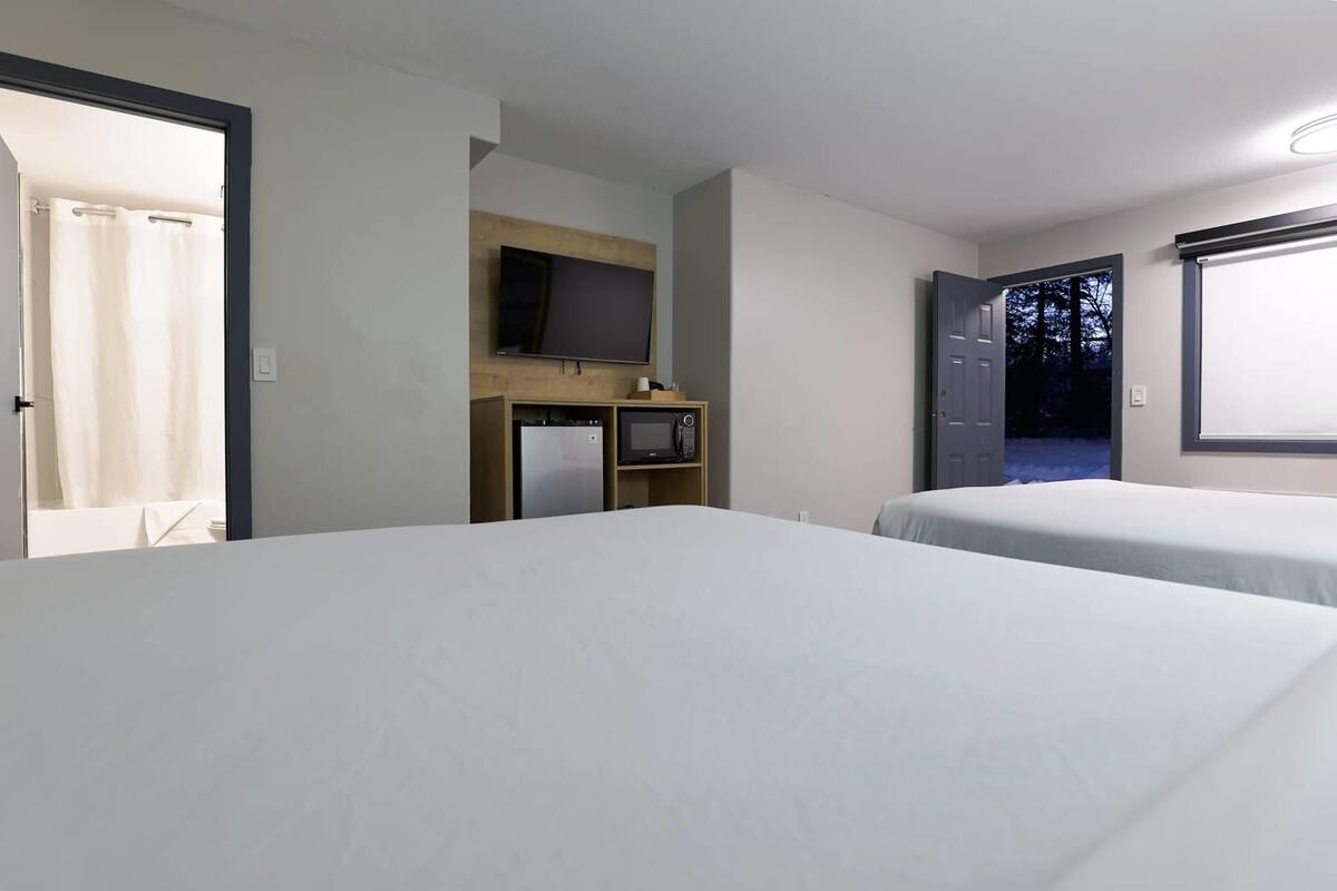 A hotel room with two beds, a television on a wooden shelf, a small refrigerator, and a window with a view of trees.