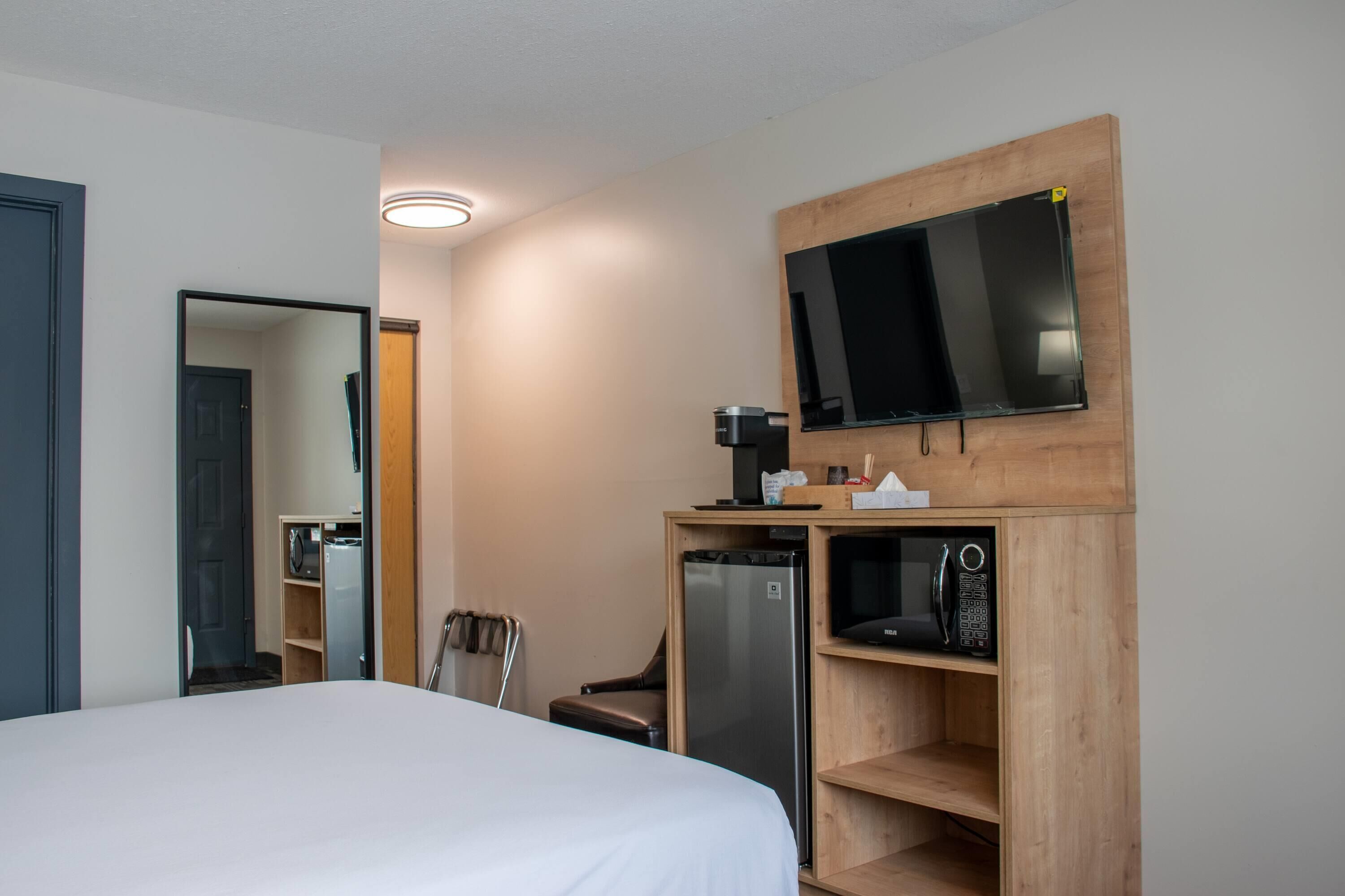 A hotel room with a bed, a television mounted on a wooden shelf, a refrigerator, and a microwave.