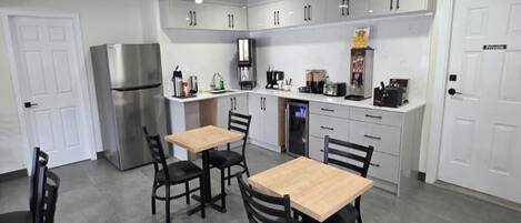 A modern kitchen with white cabinets, a stainless steel refrigerator, a coffee machine, and a microwave. There are two wooden tables with chairs.