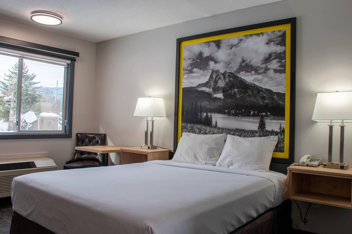 A hotel room with a bed, two bedside lamps, a nightstand with a phone, a window with a view of trees and mountains, and a large framed picture on the wall.