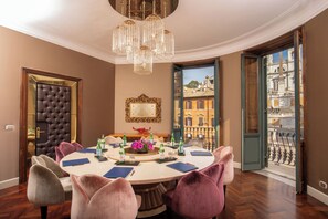 Une salle Ă manger avec une table ronde, des chaises, un lustre, un miroir et une vue sur les bĂątiments Ă travers une porte ouverte.