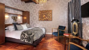 A bedroom with a bed, a chandelier, a chair, a desk, and a painting on the wall.