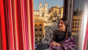 Una persona de pie junto a una ventana con vistas a un edificio histórico y una escalera.