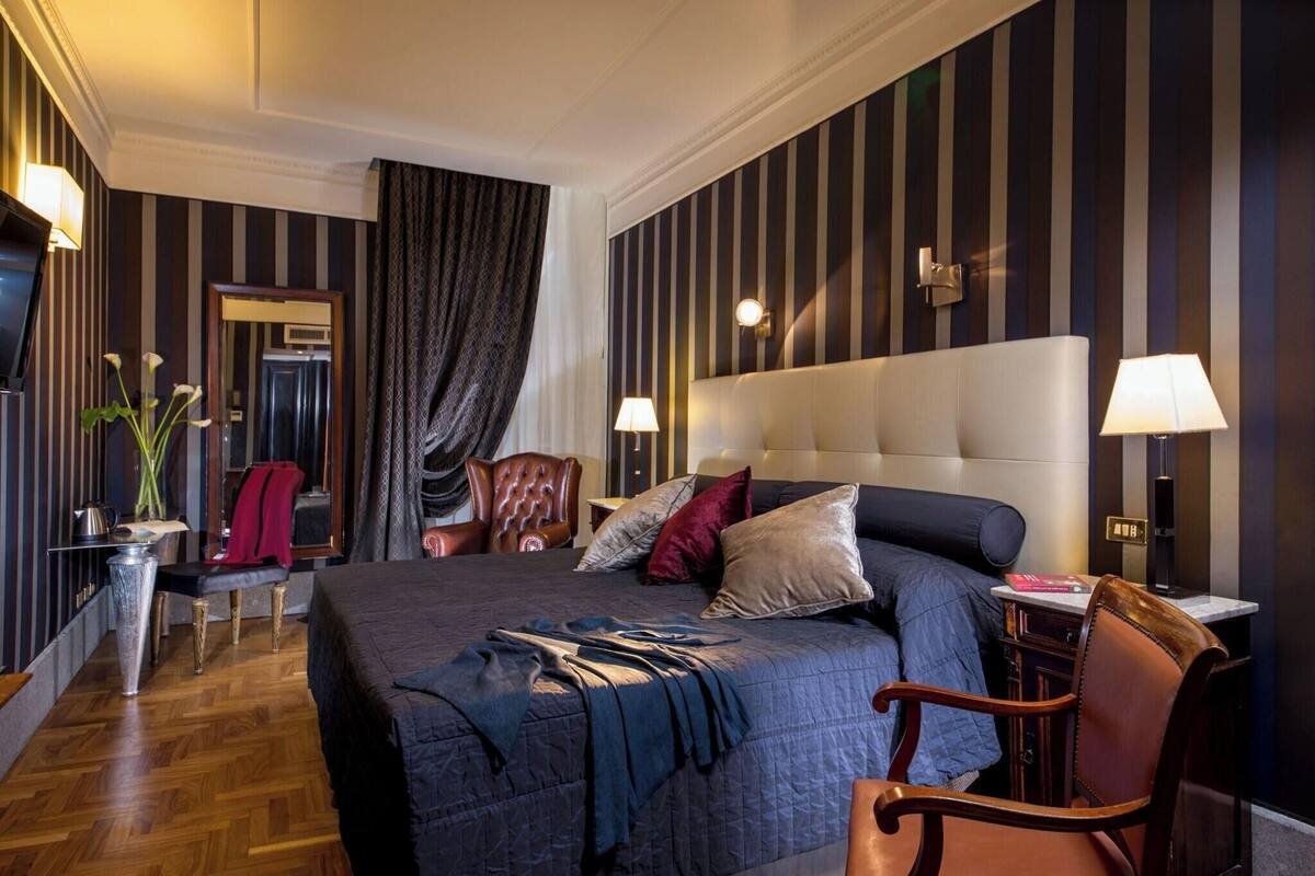 A hotel room with a large bed, two bedside lamps, a wooden chair, a small table, and a mirror.