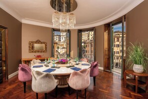A dining area with a round table, chairs, a chandelier, a mirror, and a view of buildings through open doors.