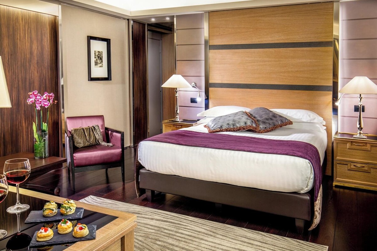 A hotel room with a bed, two bedside tables, a chair, a desk with a vase of flowers, and a small table with snacks.