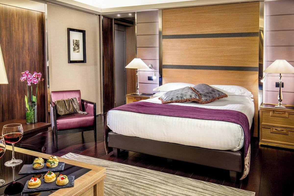 A hotel room with a bed, two bedside tables, a chair, a desk with a vase of flowers, and a small table with snacks.