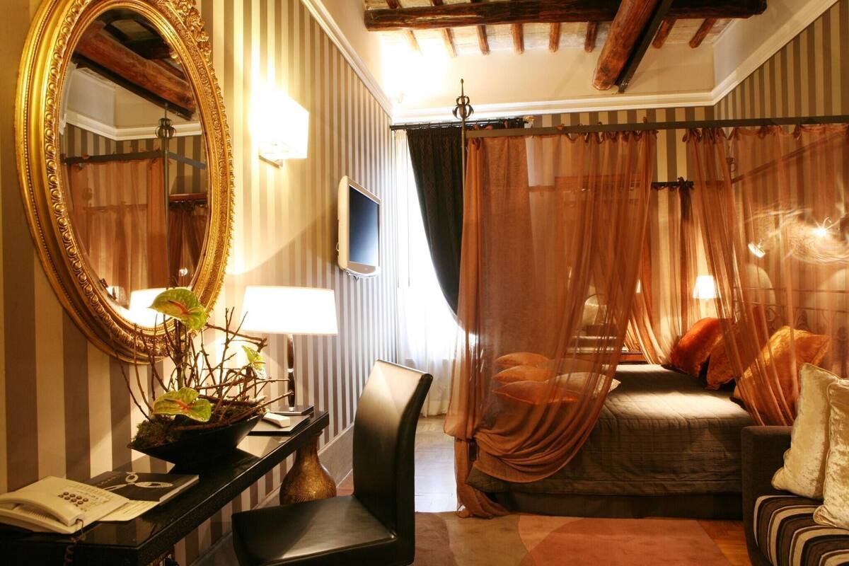A hotel room with a large mirror, a bedside table with a lamp, a chair, a bed with curtains, and a television.