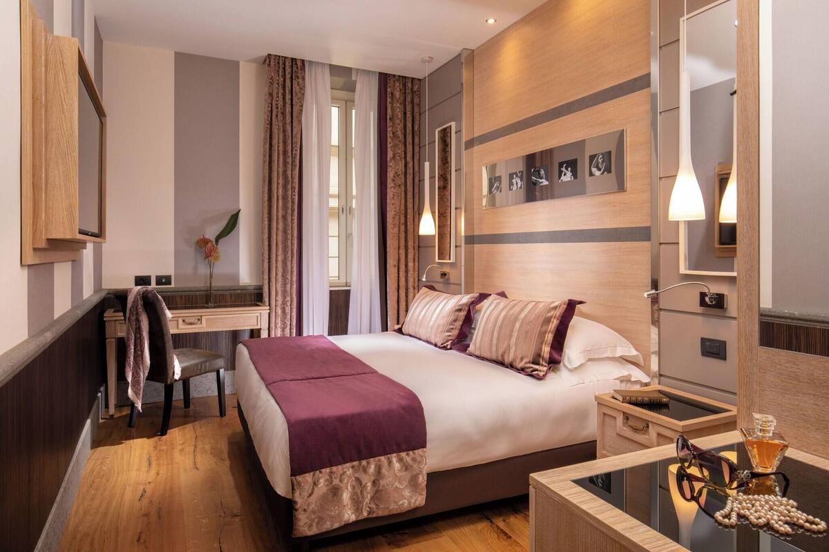 A hotel room with a bed, a desk, a chair, a mirror, and a bedside table with a lamp and a glass of water.