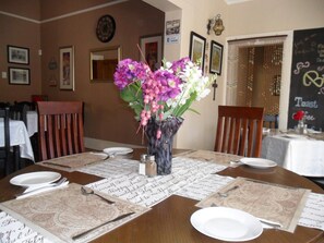 Una mesa de comedor dispuesta con platos blancos, cubiertos y un jarrón con flores moradas.