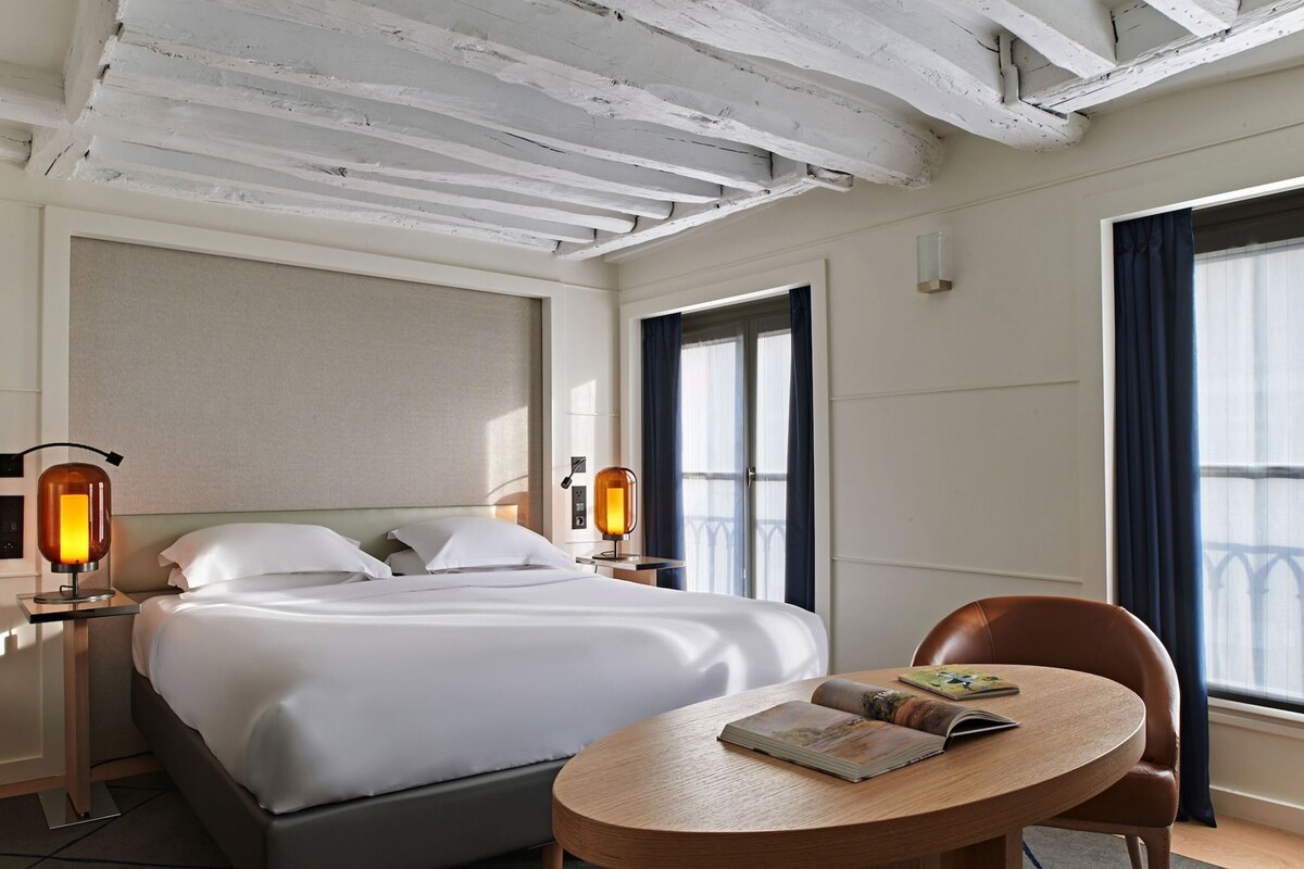 A hotel room with a bed, bedside lamps, a round table with a book, and a chair.