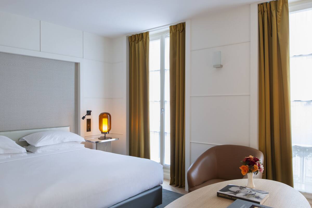 A hotel room with a bed, a nightstand with a lamp, a chair, and a table with a vase of flowers.