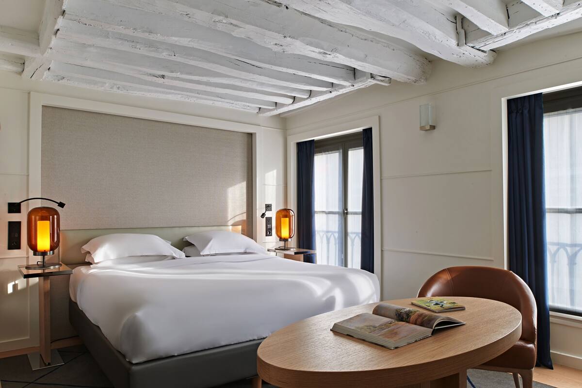 A hotel room with a bed, bedside lamps, a round table with a book, and a window with curtains.