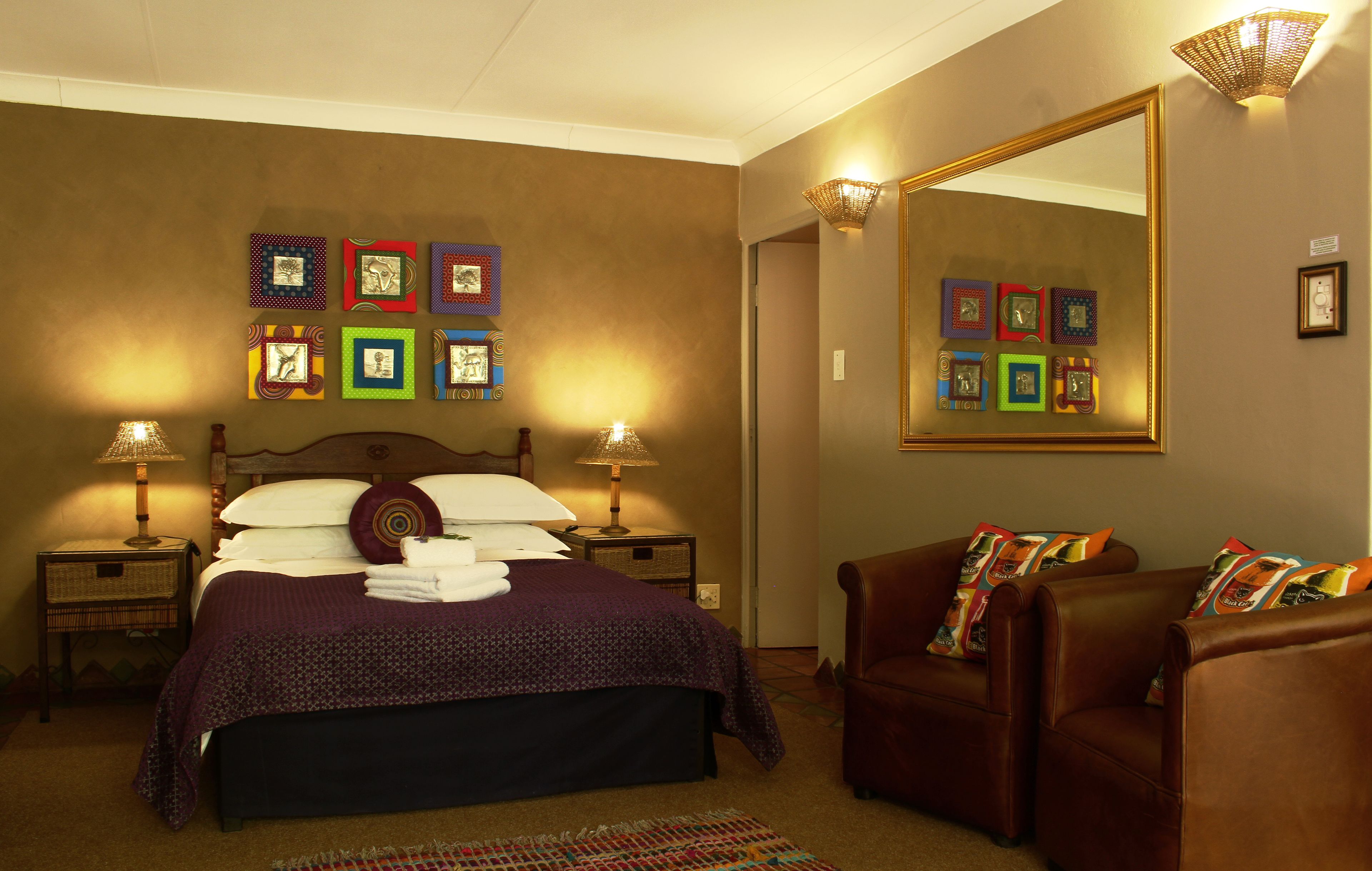 A neatly made bed with a purple bedspread and pillows, two bedside lamps, a large mirror, and a brown leather chair with colorful cushions.