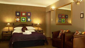 A neatly made bed with a purple bedspread and pillows, two bedside lamps, a large mirror, and a brown leather chair with colorful cushions.
