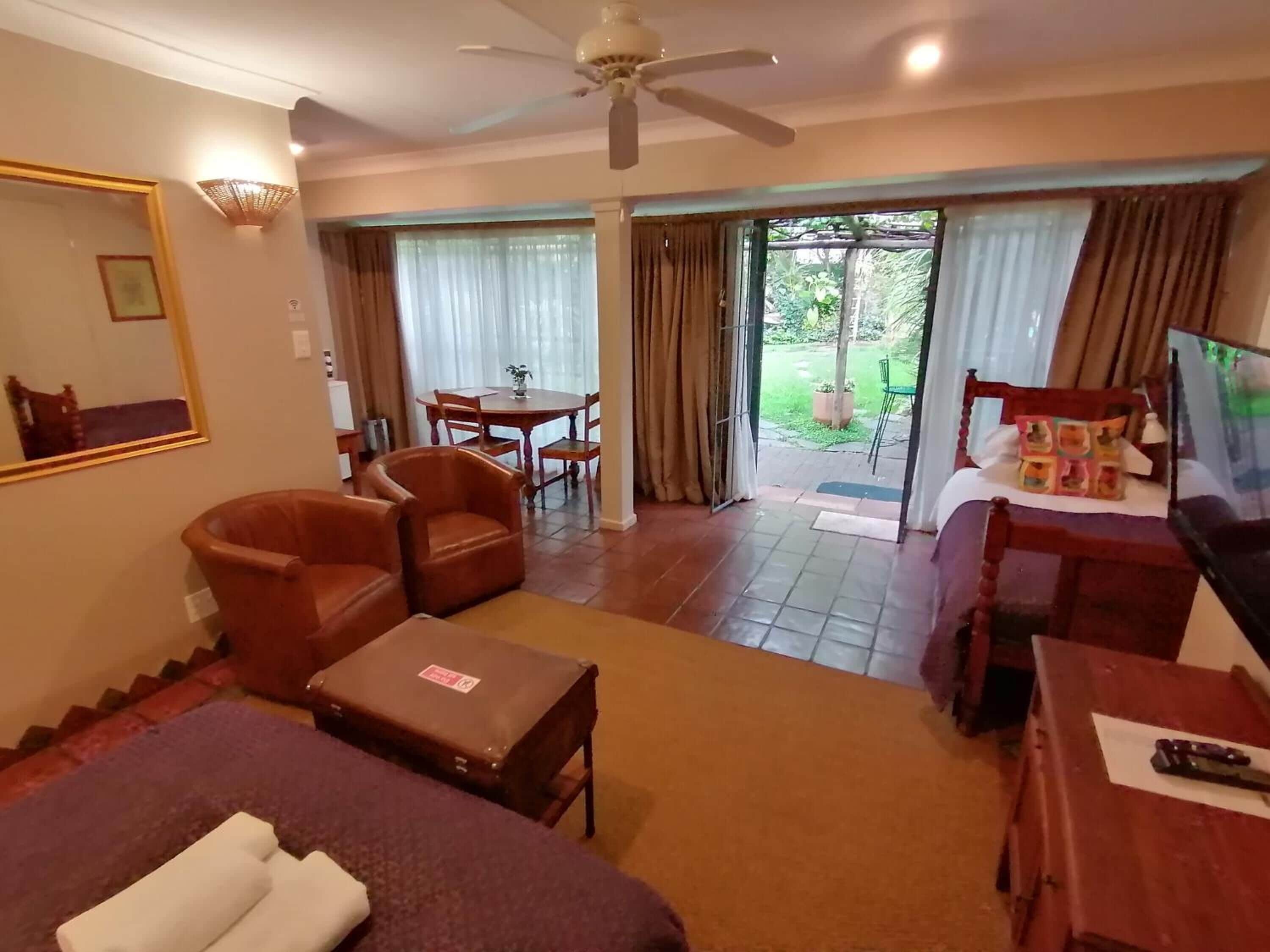 A hotel room with a bed, a desk, a chair, a table, a ceiling fan, and a view of the garden.