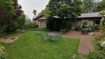 Un patio trasero bien cuidado con césped, una zona de patio con mesas y sillas, y una variedad de plantas y árboles.