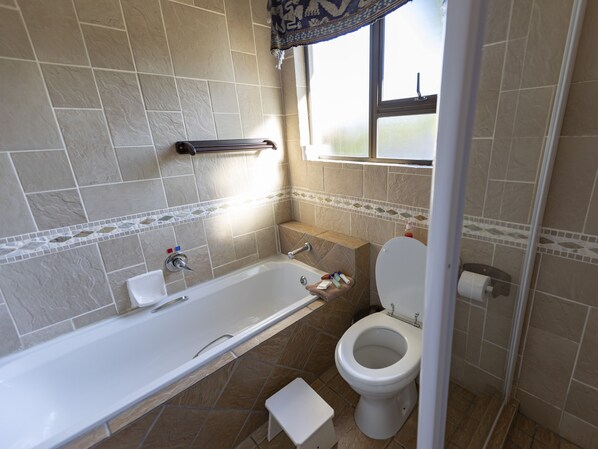 A bathroom with a bathtub, toilet, and a window. - Lombok Lodge (Port Shepstone)
