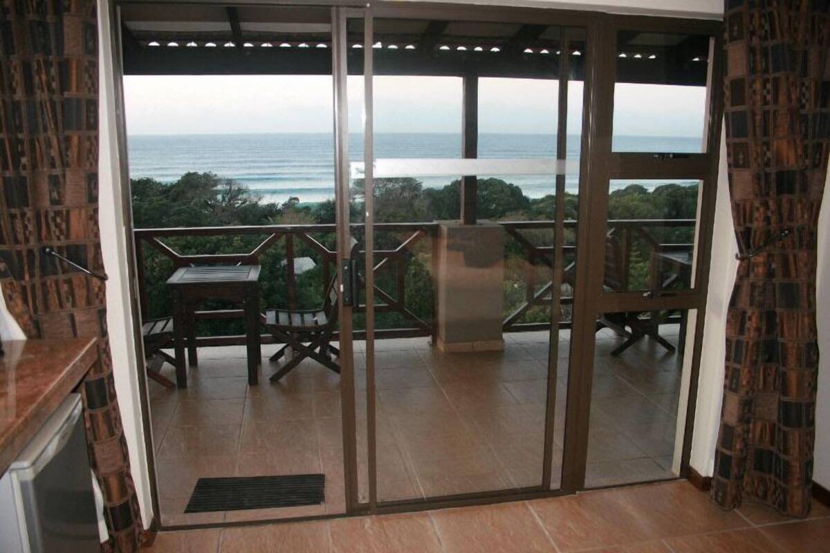 A balcony with a view of the ocean, a table, and chairs.