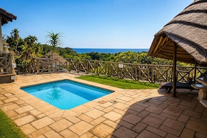 Una piscina con vista al océano, rodeada por una terraza de madera y una estructura con techo de paja.