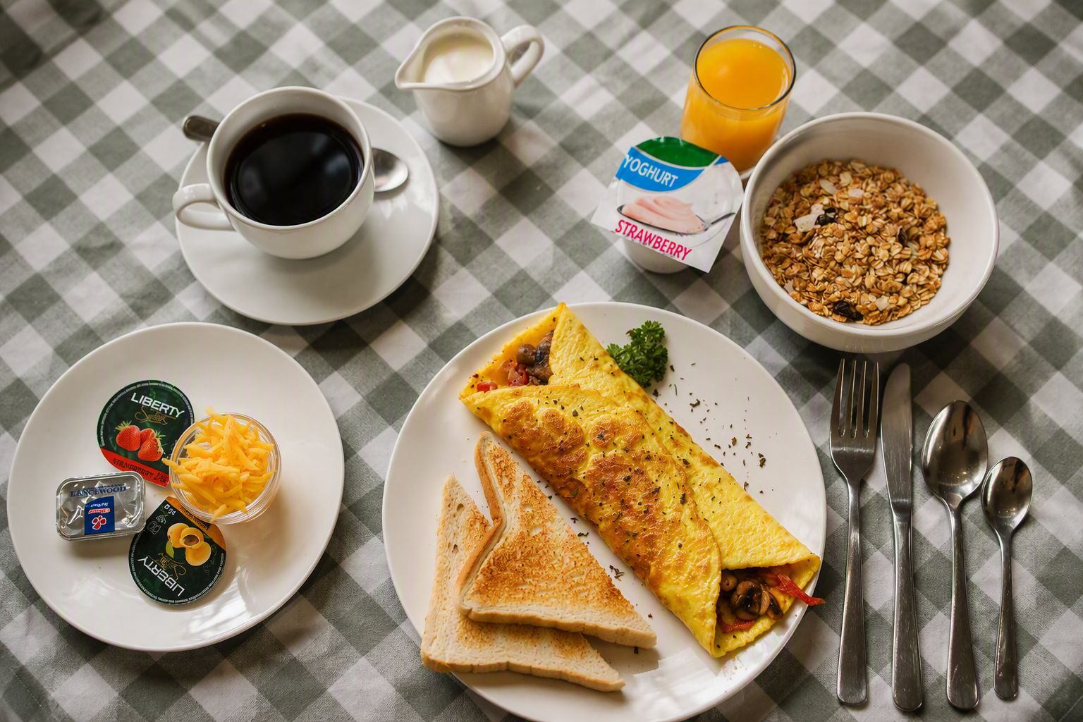 Un desayuno con tostadas, una tortilla, una taza de café, un vaso de zumo de naranja, un bol de granola y dos recipientes de yogur.