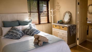 Una cama bien tendida con almohadones estampados, una mesita de noche con espejo y una pequeña cesta, y una ventana con persianas.