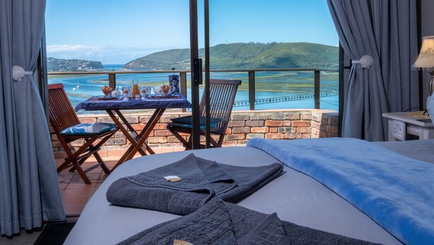Una habitación de hotel con una cama, una mesa con una botella y vasos, y vistas al mar y las colinas.