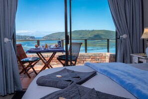 Una camera d'albergo con un letto, un tavolo con una bottiglia e dei bicchieri, e vista sul mare e sulle colline.