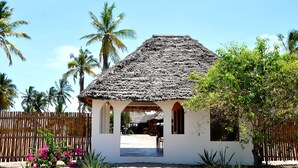 A small, thatched-roof structure with a white wall and wooden arches, surrounded by palm trees and a wooden fence. - Kijongo Bay Beach Resort (Pangani)