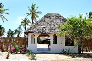 A small, thatched-roof structure with a white wall and wooden arches, surrounded by palm trees and a wooden fence. - Kijongo Bay Beach Resort (Pangani)
