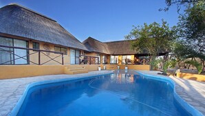 Un área de piscina con una construcción de techo de paja, escaleras que conducen a un espacio cubierto para sentarse y un cielo azul despejado.