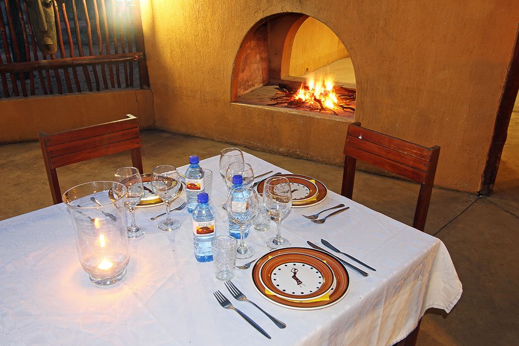 Zona de comedor al aire libre con una mesa preparada para dos, con mantel blanco, platos, cubiertos y vasos. Una pequeña vela está encendida sobre la mesa, y hay botellas de agua y una botella de refresco. Al fondo, se observa un horno de ladrillo con fuego encendido en su interior.