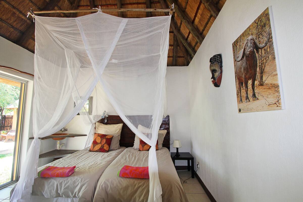 Una habitación con cama bajo dosel, una mesita de noche con lámpara, un cuadro de un elefante en la pared y una ventana con vistas al exterior.