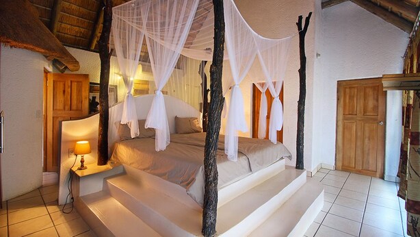 Una cama con dosel con sábanas blancas, una mesita de noche con lámpara y una puerta de madera en una habitación con paredes blancas y suelo de baldosas.