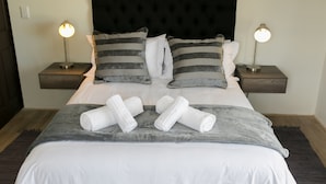 A neatly made bed with white linens, grey pillows, and rolled towels. There are two bedside lamps on nightstands. - 401 Rozendal Cottages (Stellenbosch)