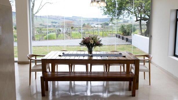 A dining area with a wooden table and chairs, a vase with flowers, and a large window offering a view of a vineyard and landscape. - 401 Rozendal Cottages (Stellenbosch)