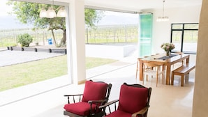 A spacious room with a large window offering a view of a vineyard. There are two red upholstered chairs and a wooden dining table with benches. - 401 Rozendal Cottages (Stellenbosch)
